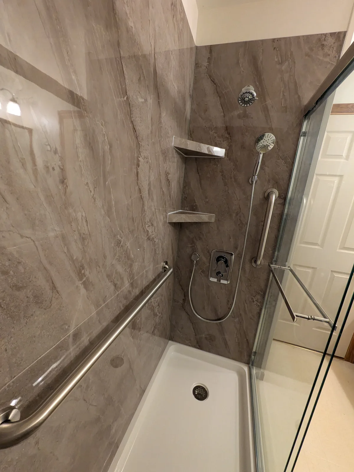 Walk-in shower in grey marble-look panels with grab bars, corner shelves and a glass door