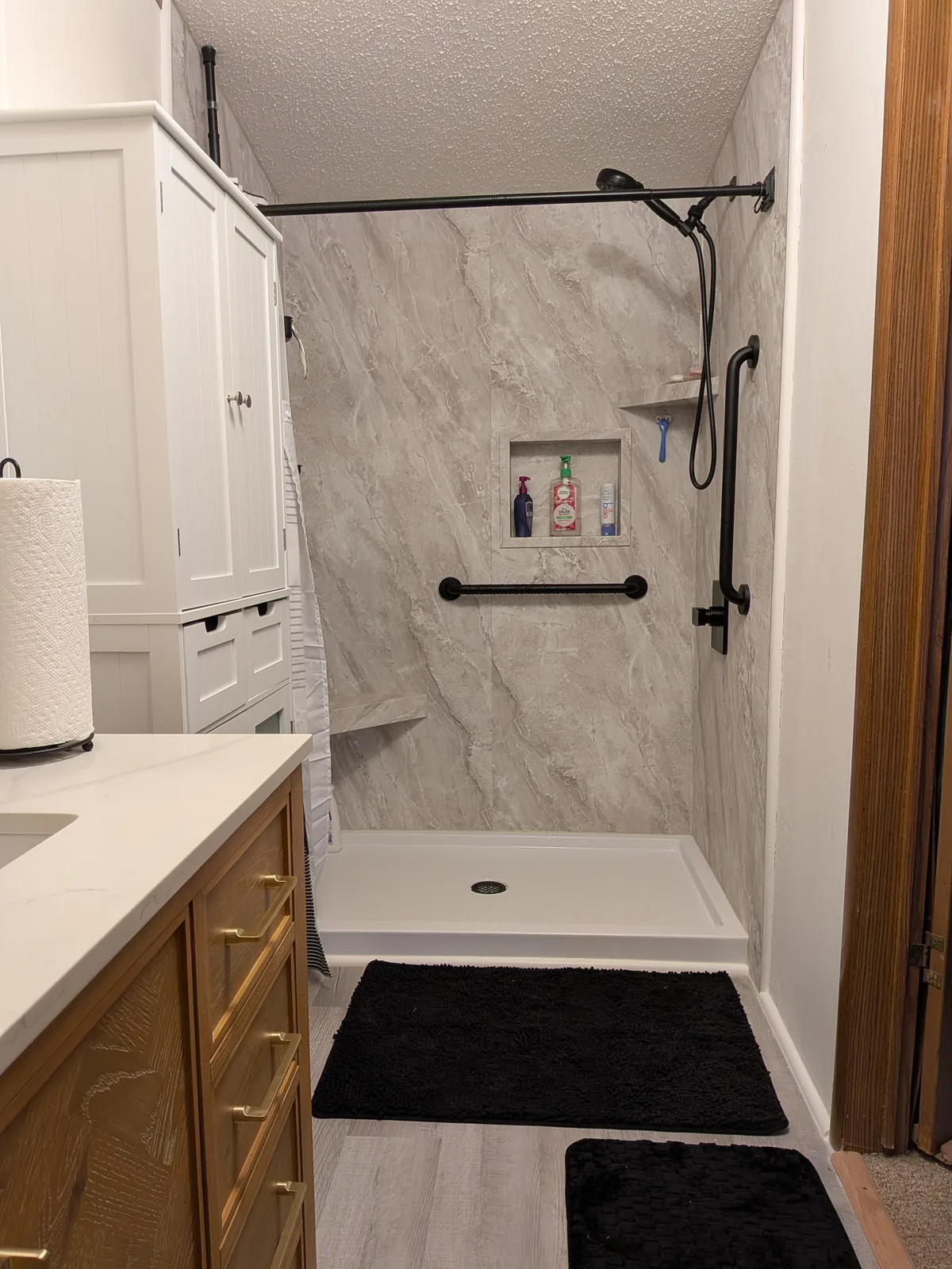 Finished walk-in shower in marble-look panels beside a wood vanity with a white quartz top