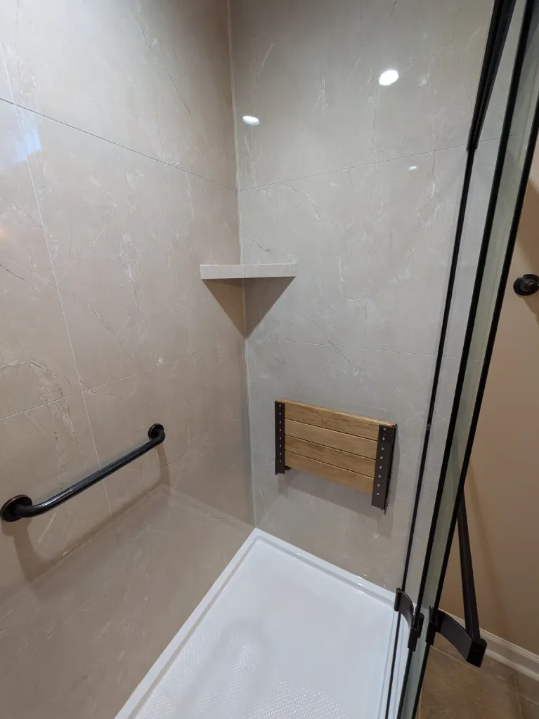 Fold-down teak bench and bronze grab bar mounted into solid backing, beside a corner shelf in the tiled wall