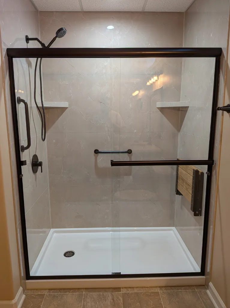 The same wall after work: a walk-in shower in marble-look tile behind bronze sliding doors, with a low-threshold base and a fold-down teak bench