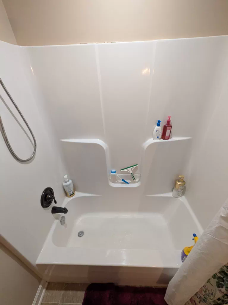 The bathroom before work: a white one-piece tub and shower surround with moulded shelves, against beige walls